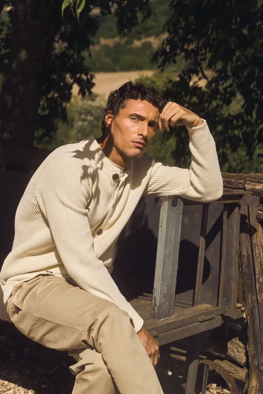 Uomo in cardigan color crema seduto su un carretto di legno, tra gli alberi