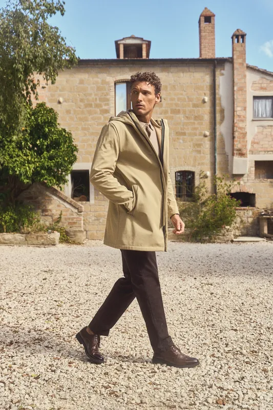 Uomo con cappotto beige che cammina nel cortile di ghiaia di un casale