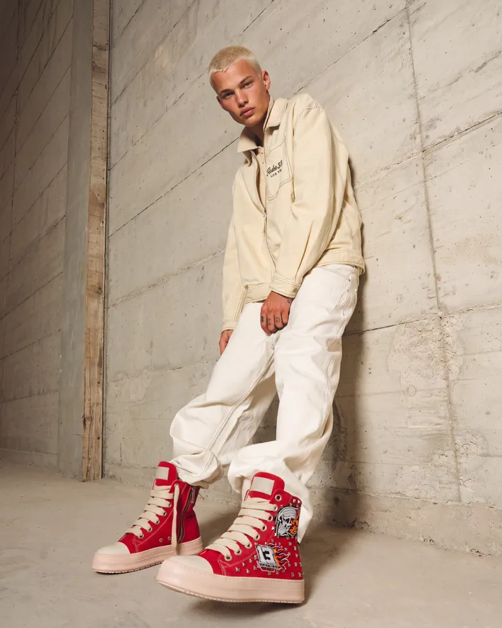 Ragazzo con felpa e pantaloni color crema e sneakers rosse, appoggiato a un muro di cemento