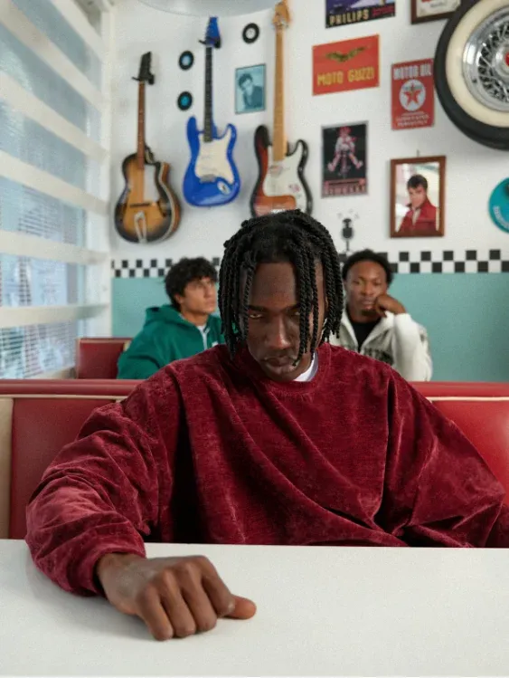 Ragazzo con maglione bordeaux al tavolo, con chitarre appese alle pareti